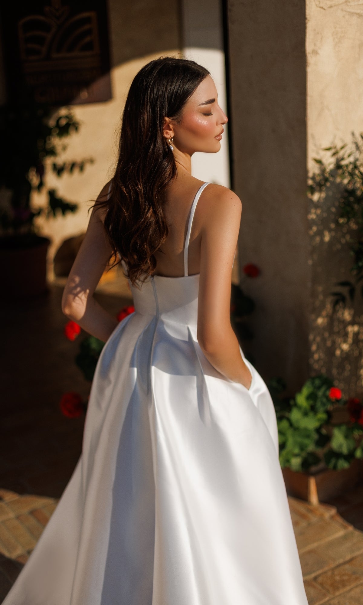 A woman in the Classic Long White Formal Dress: Tina Holly TE305 stands outdoors, hands in pockets, facing away from the camera.